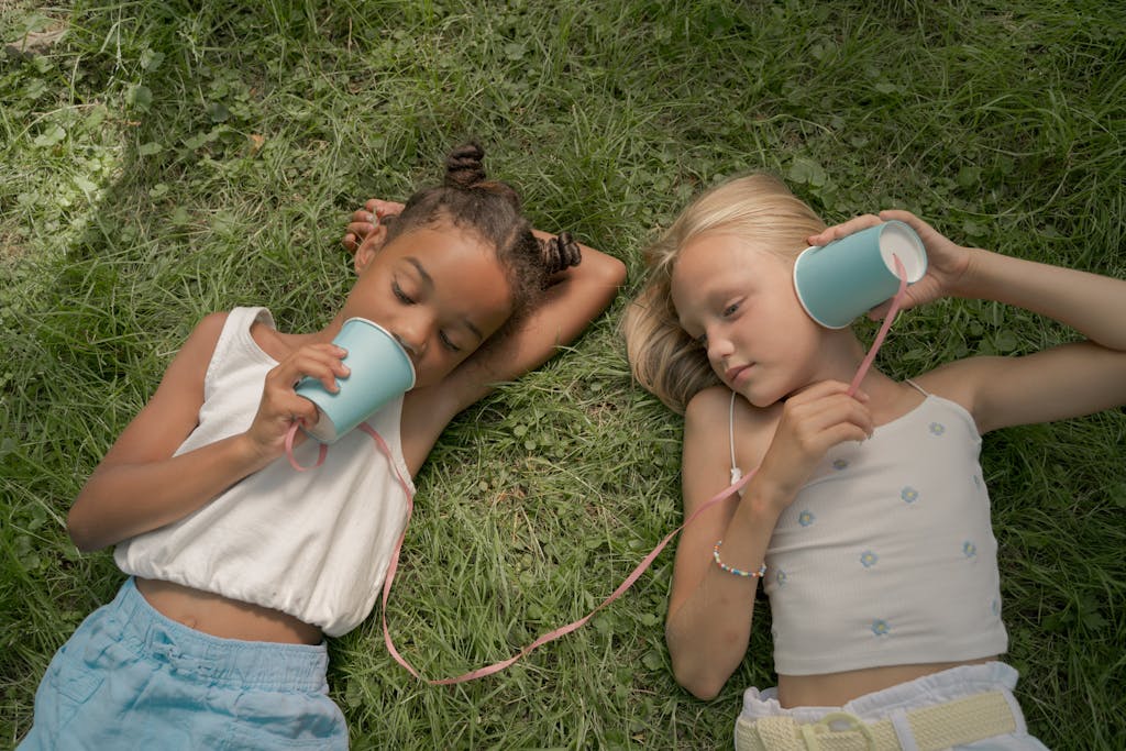Two Teenage Girls Laying on Grass and Playing Telephone Call Using Paper Cups on String